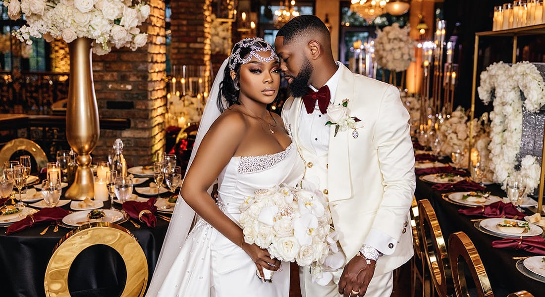 Black Bride and Groom in Luxurious Wedding Portrait | Gold Accent Reception with White Rose Details and Bouquet | Bride in Crystal Head Piece and Long Veil, Fitted Wedding Dress with Bedazzled Detailing | Groom in White Tux and Burgundy Tie
